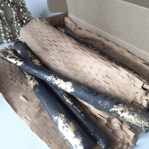 May include: A set of black taper candles with gold leaf accents, presented in a cardboard box with brown paper packaging. The candles have a textured appearance and are designed for decorative use.