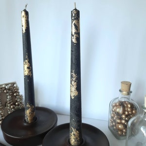 May include: Two black taper candles with gold leaf accents stand in dark brown candle holders. The candles are unlit and have a rustic, textured appearance. Decorative items, including glass bottles, are in the background.
