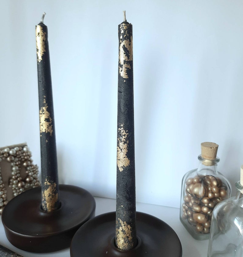 May include: Two black taper candles with gold leaf accents stand in dark brown candle holders. The candles are unlit and have a rustic, textured appearance. Decorative items, including glass bottles, are in the background.