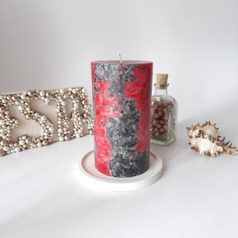 May include: A red and black pillar candle with a speckled finish sits on a white pedestal. The candle is on a white surface.