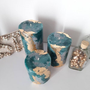 Puede incluir: Tres velas de pilar color turquesa con detalles de pan de oro. Las velas var&iacute;an en altura y est&aacute;n dispuestas juntas. Un collar de perlas y un tarro de cristal con cuentas doradas est&aacute;n en el fondo.