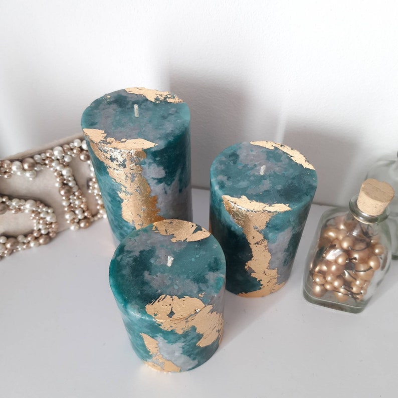 Puede incluir: Tres velas de pilar color turquesa con detalles de pan de oro. Las velas var&iacute;an en altura y est&aacute;n dispuestas juntas. Un collar de perlas y un tarro de cristal con cuentas doradas est&aacute;n en el fondo.