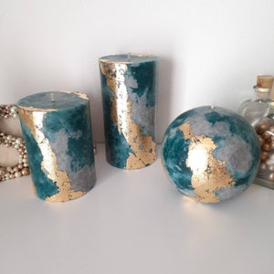 Puede incluir: Tres velas decorativas de diferentes formas: un cilindro, un cilindro m&aacute;s alto y una esfera. Las velas son de color verde azulado y gris con detalles de pan de oro. Las velas est&aacute;n sobre una superficie blanca.