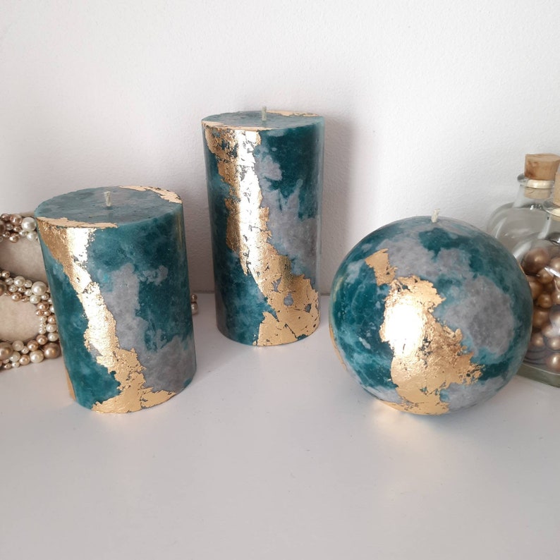 Puede incluir: Tres velas decorativas de diferentes formas: un cilindro, un cilindro m&aacute;s alto y una esfera. Las velas son de color verde azulado y gris con detalles de pan de oro. Las velas est&aacute;n sobre una superficie blanca.
