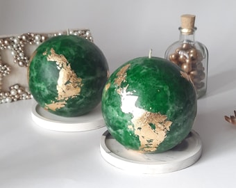 May include: Two green globe-shaped candles with gold accents on white marble coasters.