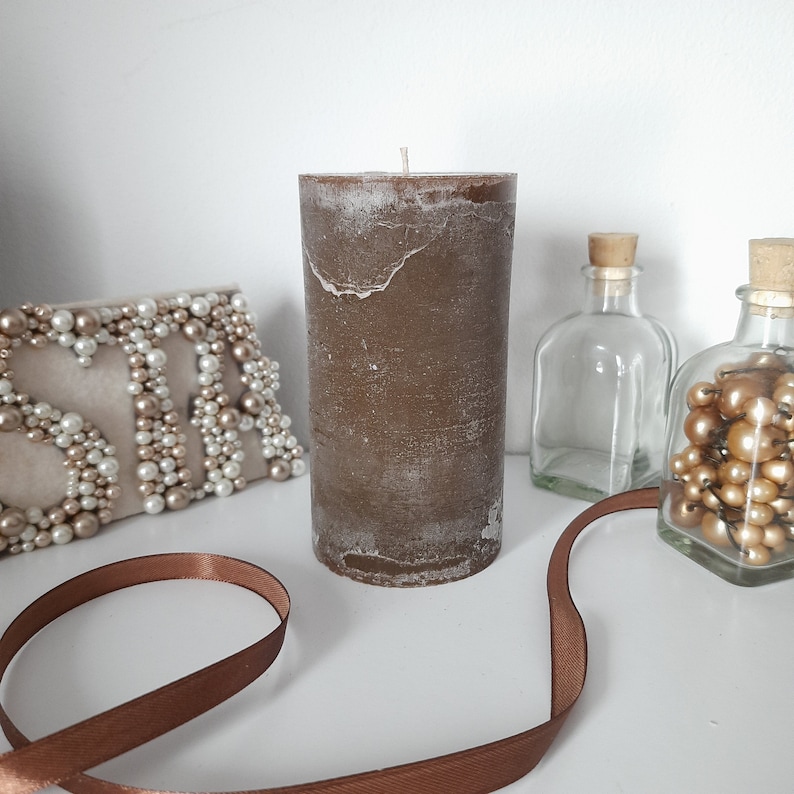 May include: A brown pillar candle with a white, textured finish. The candle is sitting on a white surface with a brown ribbon and decorative items.