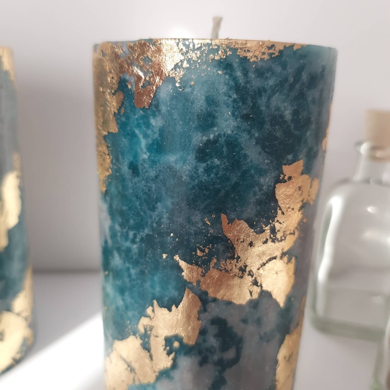 Puede incluir: Una vela de pilar de color verde azulado con detalles de pan de oro. La vela tiene un aspecto veteado y una sola mecha. Una botella de vidrio transparente es visible en el fondo.