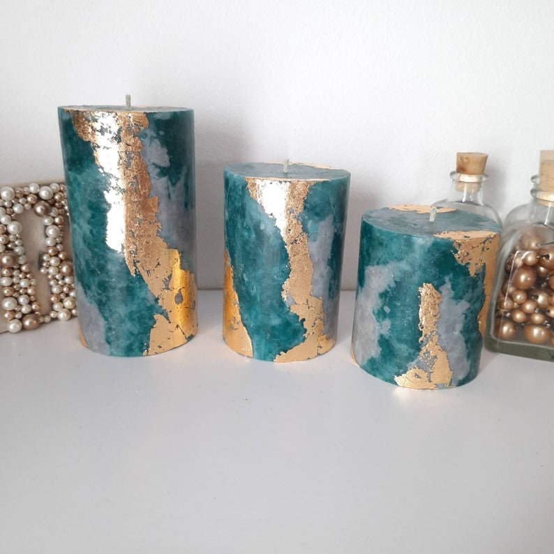 Puede incluir: Tres velas de pilar decorativas de diferentes alturas. Las velas son de color verde azulado, gris y con dise&ntilde;o de pan de oro. Las velas cil&iacute;ndricas tienen un aspecto marmolado. Los detalles de pan de oro a&ntilde;aden un toque de elegancia. Cada vela tiene una mecha.