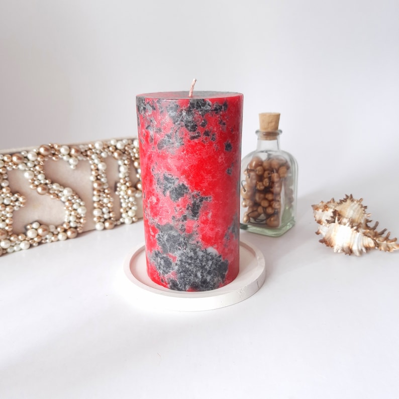 May include: A red and black speckled candle on a white pedestal. The candle is in a cylindrical shape and has a black wick.