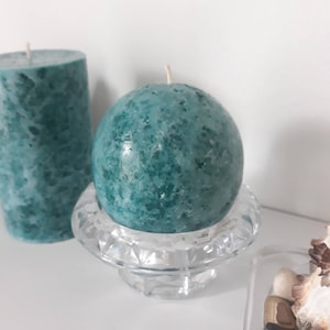 May include: Two teal blue candles, one round and one cylindrical, on a clear glass candle holder. The round candle is on the holder, and the cylindrical candle is on a white surface.