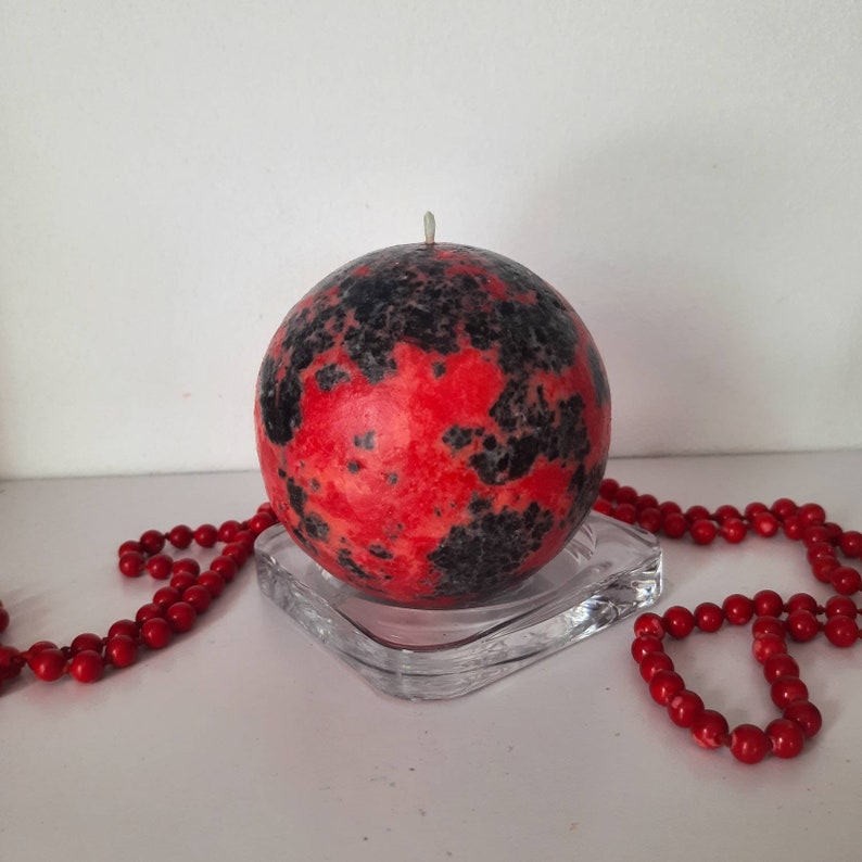 May include: A red and black speckled sphere candle on a clear glass base. The candle is sitting on a white surface with a red beaded necklace.