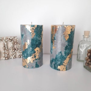 Puede incluir: Dos velas cil&iacute;ndricas con un dise&ntilde;o de color turquesa, gris y pan de oro. Las velas tienen un aspecto veteado con detalles de pan de oro. Las velas est&aacute;n sobre una superficie blanca, con objetos decorativos en el fondo.