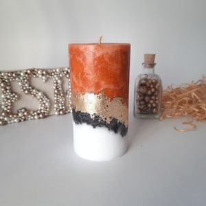 Puede incluir: Una vela de pilar decorativa con un diseño en capas. La sección superior es naranja, seguida de pan de oro, negro y blanco en la base. La vela tiene una sola mecha y mide aproximadamente 10 cm de alto.