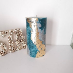 May include: A teal and grey marbled candle with gold leaf detailing. The cylindrical candle has a single wick and is displayed on a white surface. This decorative candle would make a lovely home decor item.