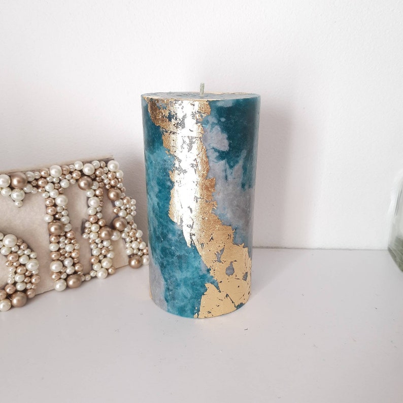Puede incluir: Una vela cil&iacute;ndrica con un dise&ntilde;o veteado en color turquesa y gris, acentuada con pan de oro. La vela tiene una sola mecha y se exhibe sobre una superficie blanca. Un elemento decorativo para el hogar.