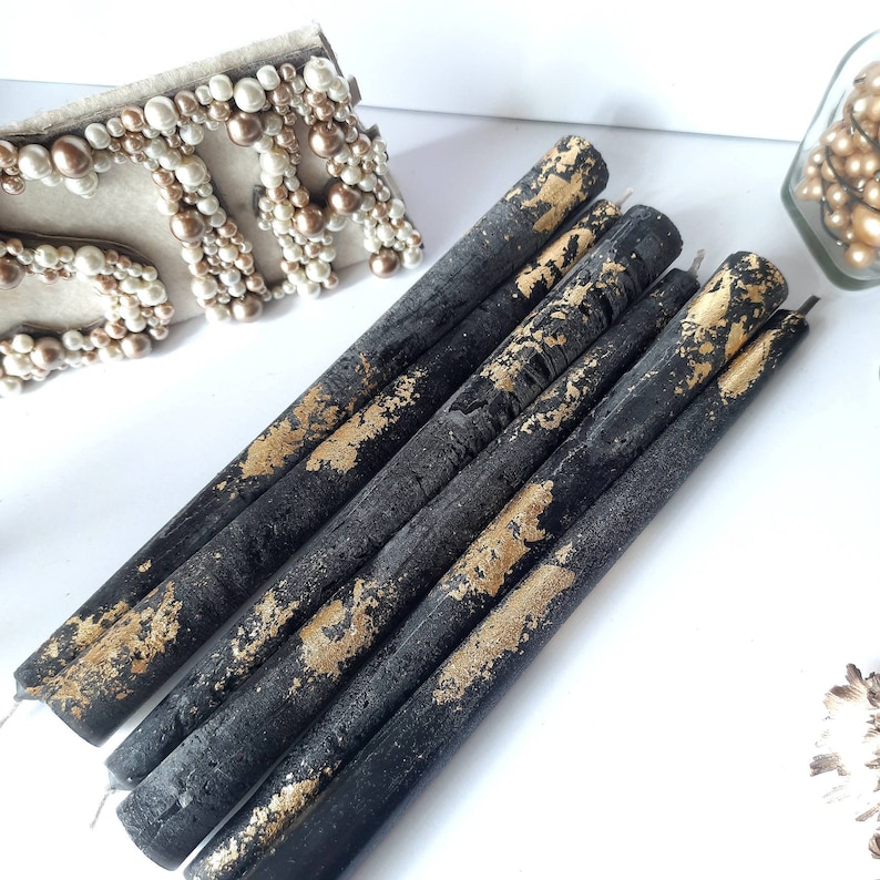 May include: A collection of black taper candles with gold leaf accents. The candles are arranged on a white surface, with a decorative beaded item and a glass jar with pearls in the background. The candles have a rustic, textured appearance.