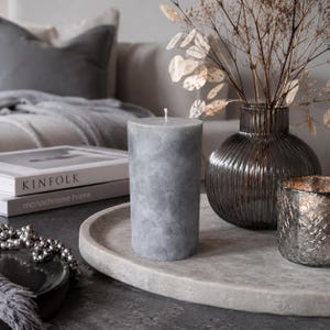 Può includere: Una candela cilindrica a colonna di colore grigio con una texture marmorizzata, posizionata su un vassoio rotondo in pietra. La candela presenta uno stoppino bianco singolo al centro. La superficie mostra sottili variazioni di tonalità, creando un aspetto morbido e nuvoloso.