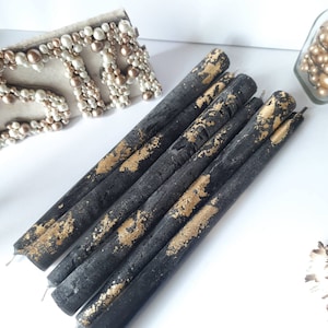 May include: A collection of black taper candles with gold leaf accents. The candles are arranged diagonally on a white surface. The gold leaf adds a touch of elegance to the dark candles, creating a striking contrast. Decorative candles for home decor.