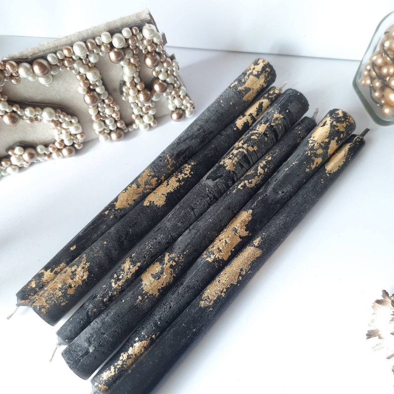 May include: A collection of black taper candles with gold leaf accents. The candles are arranged diagonally on a white surface. The gold leaf adds a touch of elegance to the dark candles, creating a striking contrast. Decorative candles for home decor.