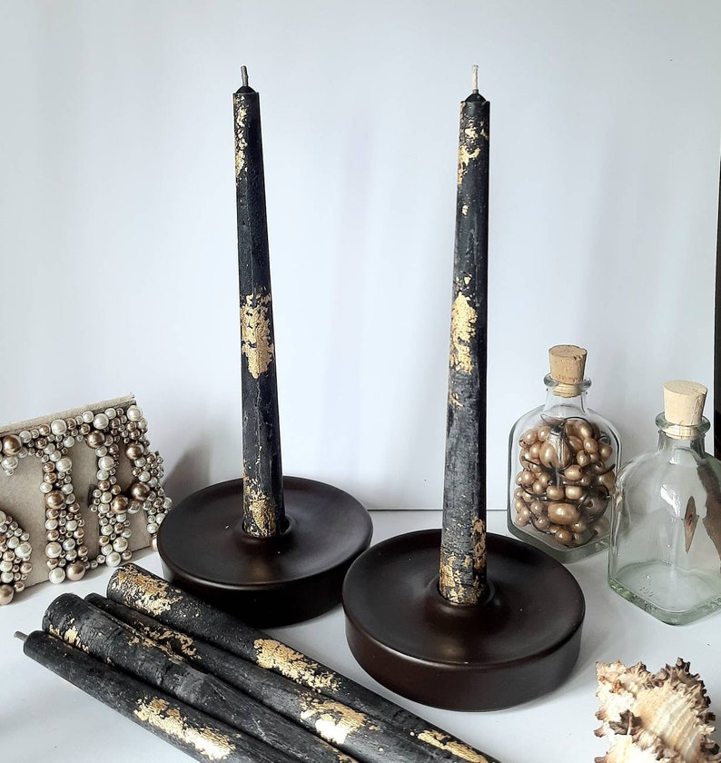 May include: Two black taper candles with gold leaf accents stand in dark brown candle holders. Several additional candles and decorative items, including small glass bottles and a beaded frame, are also visible.