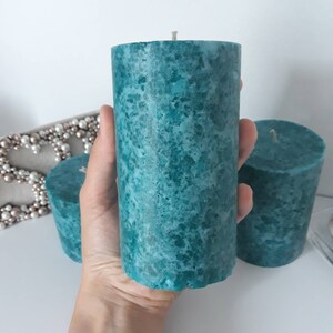 May include: Three teal green pillar candles with a speckled texture. The candles are cylindrical and have a white wick.