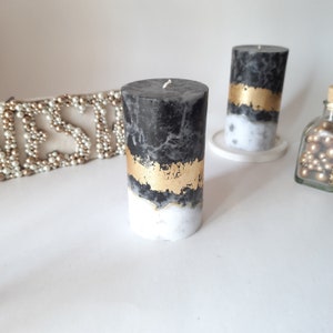 Puede incluir: Dos velas cil&iacute;ndricas con un acabado de cera veteada en negro y blanco. Cada vela est&aacute; decorada con una banda horizontal de pan de oro texturizado en el centro, que separa la secci&oacute;n superior oscura de la base blanca.