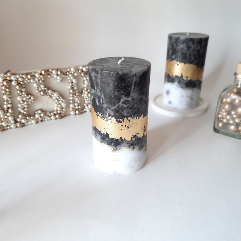 Puede incluir: Dos velas cil&iacute;ndricas con un acabado de cera veteada en negro y blanco. Cada vela est&aacute; decorada con una banda horizontal de pan de oro texturizado en el centro, que separa la secci&oacute;n superior oscura de la base blanca.