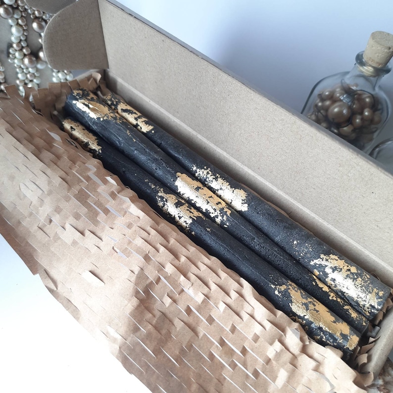 May include: A set of four black taper candles with gold leaf accents, presented in a cardboard box with brown paper packaging. The candles are long and slender, with a rustic, textured appearance.