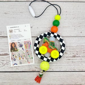 May include: A handmade car-themed teething toy with a black and white checkered ring, orange, green, and yellow beads, and a red tassel. The toy features a small orange car with yellow wheels and a green alligator design.