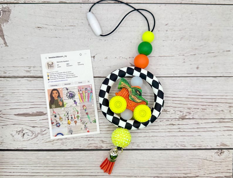 May include: A handmade car-themed teething toy with a black and white checkered ring, orange, green, and yellow beads, and a red tassel. The toy features a small orange car with yellow wheels and a green alligator design.