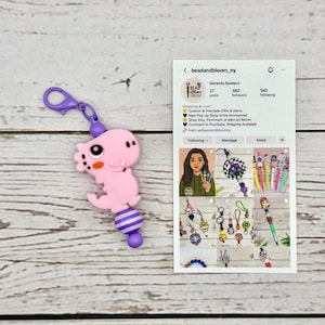 Puede incluir: Un llavero de dinosaurio rosa con un cierre morado y un acento de cuentas a rayas. El llavero está sujeto a una superficie de madera blanca. Una tarjeta de perfil de redes sociales es visible en el fondo.