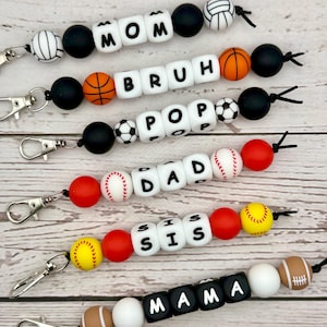 May include: A collection of beaded keychains with sports-themed and family-themed designs. Each keychain features lettered beads spelling out words like "MOM," "DAD," and "SIS," interspersed with sports ball beads such as baseballs and footballs.