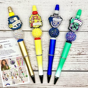 May include: Four decorative pens with themed toppers. Each pen features a different design: Hufflepuff, Gryffindor, Ravenclaw, and Slytherin. The pens have a jeweled ball and a themed charm. A business card with social media information is also visible.