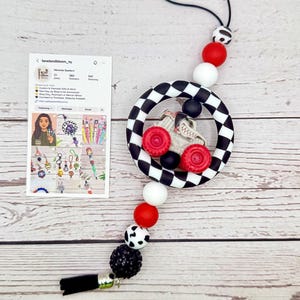 May include: A black and white checkered circle pendant with a gray race car and red wheels. The pendant is strung with red, white, black, and patterned beads, and a black tassel. A card with social media information is also present.