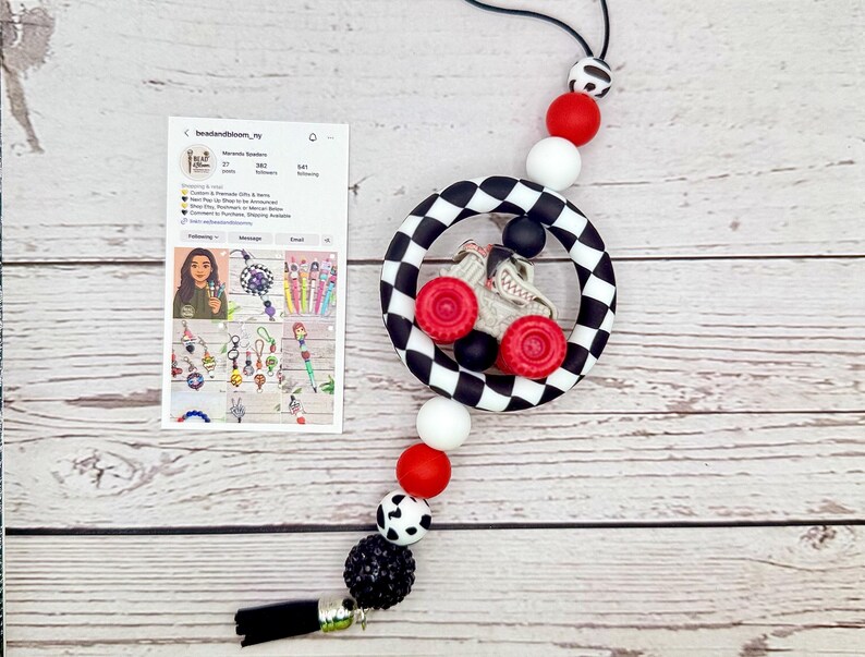 May include: A black and white checkered circle pendant with a gray race car and red wheels. The pendant is strung with red, white, black, and patterned beads, and a black tassel. A card with social media information is also present.
