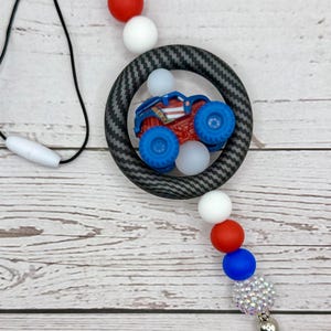 K&ouml;nnte beinhalten: Ein Schl&uuml;sselband mit einem rot, wei&szlig; und blau Thema. Das Schl&uuml;sselband hat eine schwarze Kordel, einen kreisf&ouml;rmigen Anh&auml;nger mit einem Monstertruck und eine Reihe bunter Perlen. Eine rote Quaste h&auml;ngt von unten.