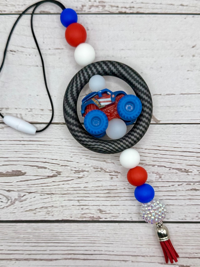K&ouml;nnte beinhalten: Ein Schl&uuml;sselband mit einem rot, wei&szlig; und blau Thema. Das Schl&uuml;sselband hat eine schwarze Kordel, einen kreisf&ouml;rmigen Anh&auml;nger mit einem Monstertruck und eine Reihe bunter Perlen. Eine rote Quaste h&auml;ngt von unten.
