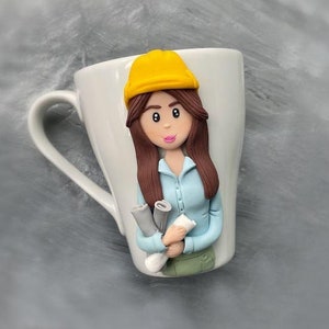 May include: White ceramic mug with a hand-painted figurine of a woman wearing a yellow hard hat and holding blueprints. The woman has brown hair and is wearing a blue shirt and green pants.