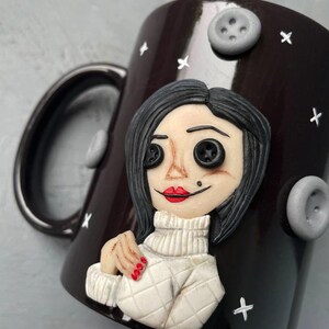 May include: A black ceramic mug with a white and black illustration of a woman with red lips and button eyes. The mug has a white handle and is decorated with white stars and grey buttons.