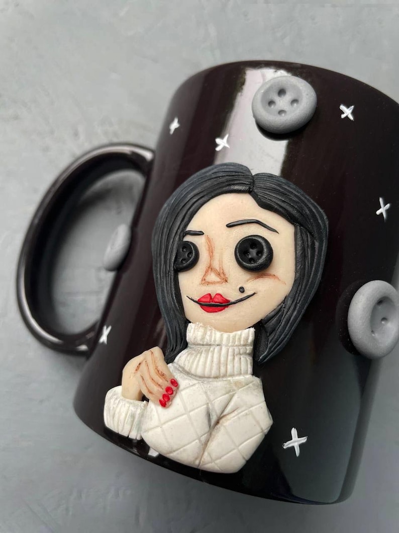 May include: A black ceramic mug with a white and black illustration of a woman with red lips and button eyes. The mug has a white handle and is decorated with white stars and grey buttons.
