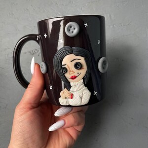 May include: A black ceramic mug with a white and gray 3D sculpted image of a woman with black hair and red lips. The mug has a white handle and is decorated with gray buttons.