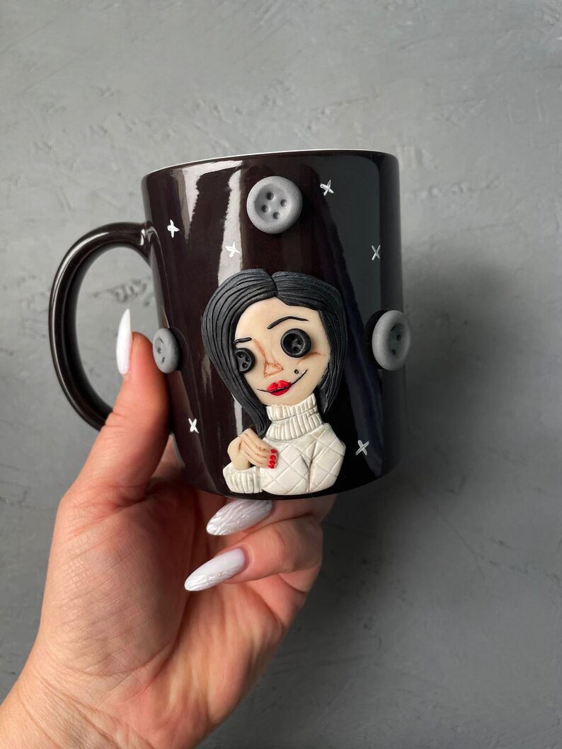 May include: A black ceramic mug with a white and gray 3D sculpted image of a woman with black hair and red lips. The mug has a white handle and is decorated with gray buttons.