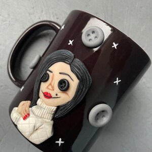 May include: A dark brown ceramic mug with a white and black illustration of a woman with buttons for eyes. The mug has a white handle and is decorated with white plus signs and gray buttons.