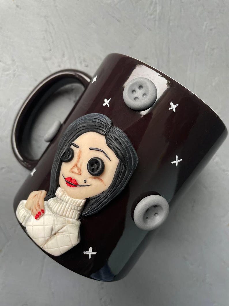 May include: A dark brown ceramic mug with a white and black illustration of a woman with buttons for eyes. The mug has a white handle and is decorated with white plus signs and gray buttons.