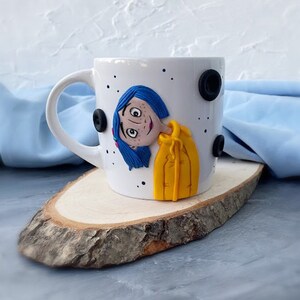 May include: White ceramic mug with a hand-painted image of a girl with blue hair and a yellow jacket. The mug has black polka dots around the rim.