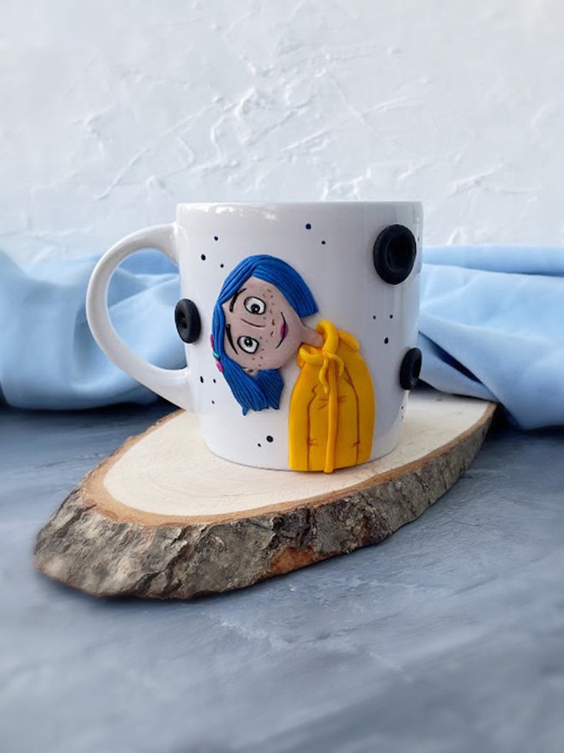 May include: White ceramic mug with a hand-painted image of a girl with blue hair and a yellow jacket. The mug has black polka dots around the rim.