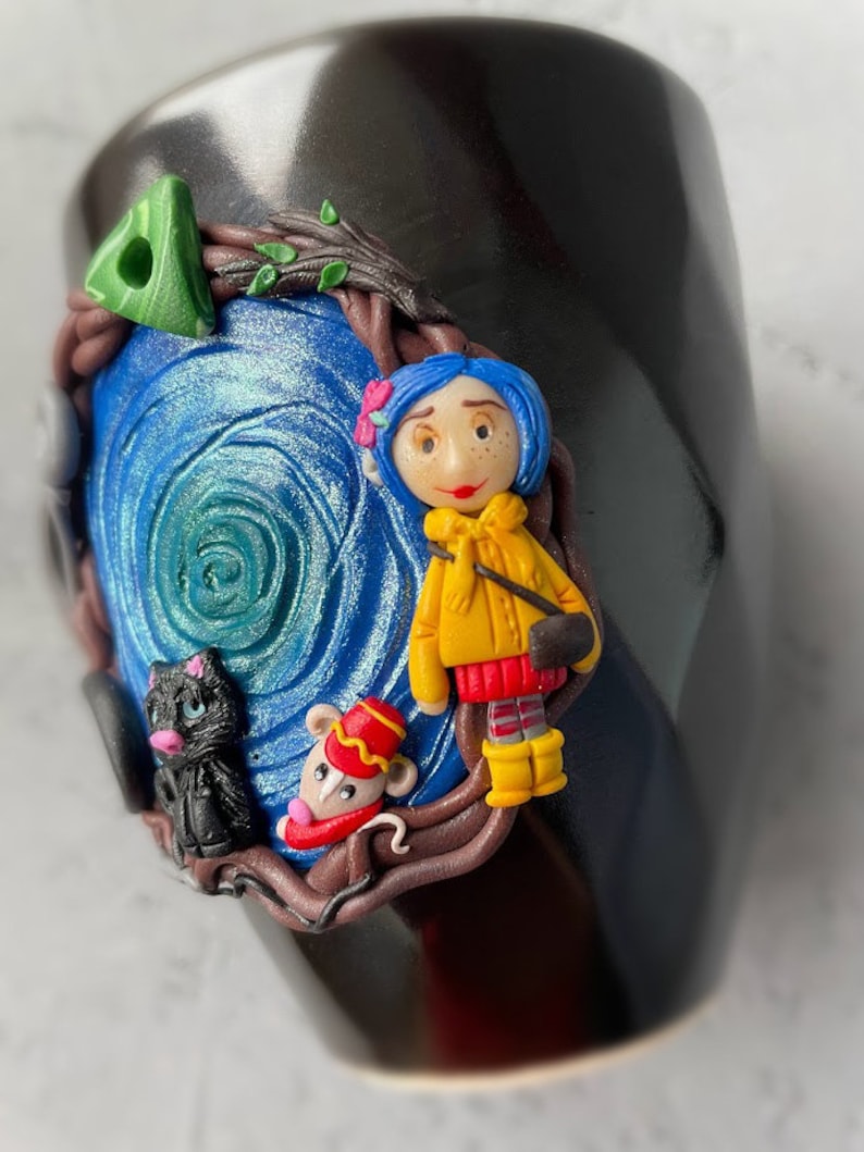 May include: Black ceramic mug with a hand-painted scene featuring a girl with blue hair, a black cat, and a mouse. The scene is set in a forest with a swirling blue background.