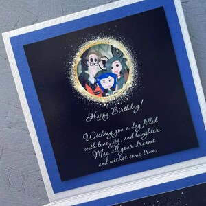 May include: A birthday card with a black background and a gold circle with a family of three characters from the movie "The Addams Family". The text on the card reads "Happy Birthday! Wishing you a day filled with love, joy, and laughter. May all your dreams and wishes come true."