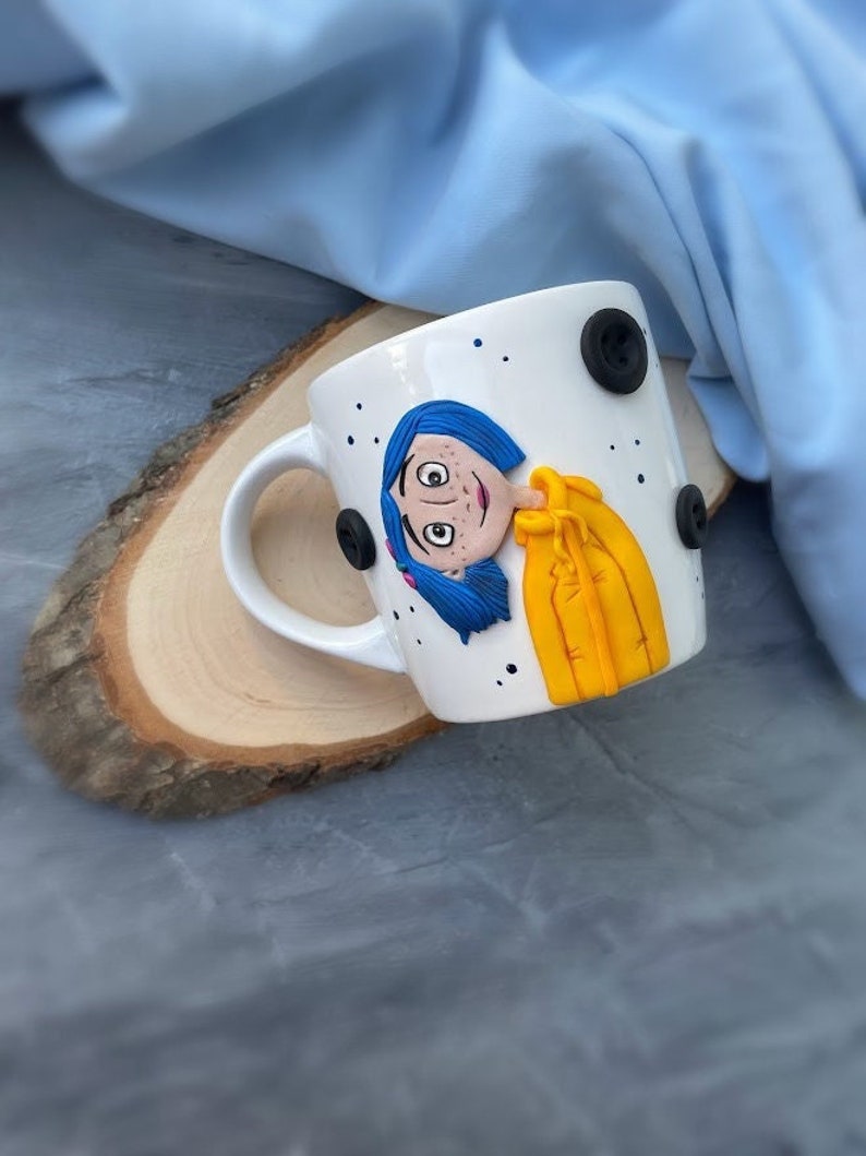 May include: White ceramic mug with a blue and yellow character design. The character has blue hair and is wearing a yellow jacket. The mug has black buttons on the side.