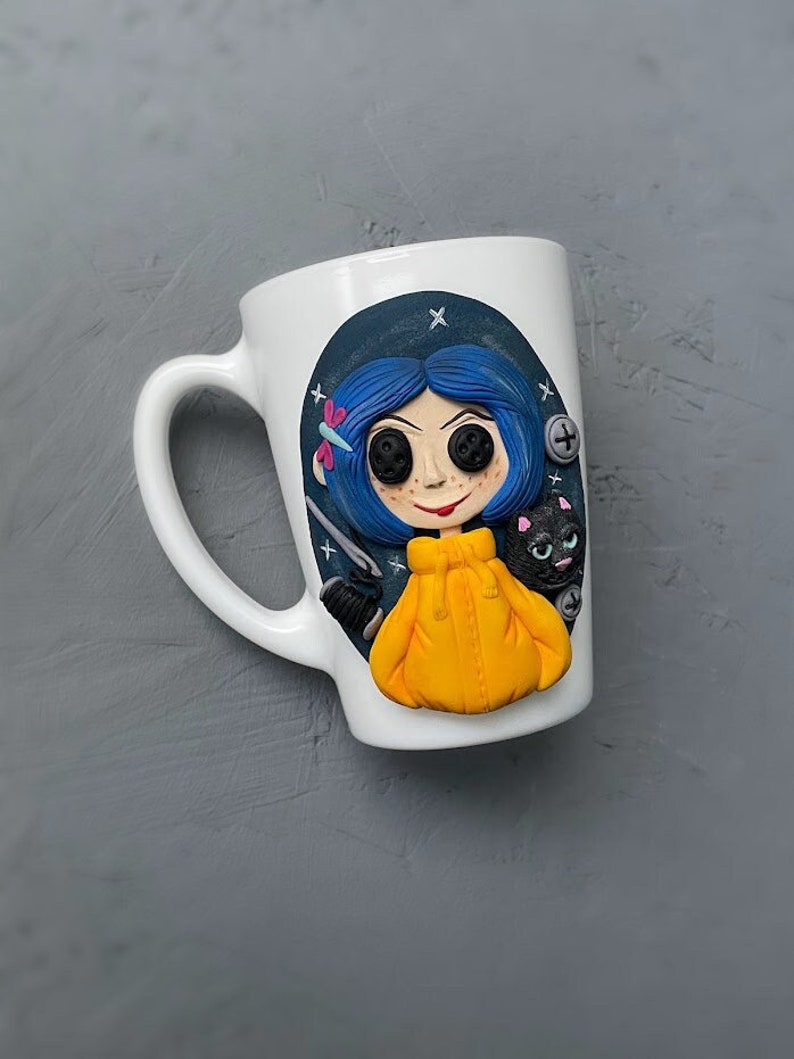 May include: White ceramic mug with a blue and yellow illustration of a girl with blue hair and a black cat. The girl is wearing a yellow jacket and has large black buttons for eyes.